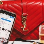 a smartphone with an Instagram profile showing on its screen beside a red YSL handbag and a bottle of Chanel perfume