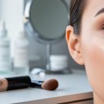 Close-up of a model’s under-eye area with blended concealer next to a concealer tube and makeup brush on a softly lit vanity.