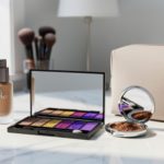 Makeup palette, foundation bottle, and compact mirror arranged on a white marble vanity with a small makeup bag in a bright, clean setting.